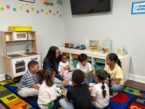 Children and two adults sitting in a circle on a colorful classroom rug during story time