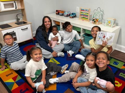 Children and two adults smiling together on a colorful classroom rug while one child holds a book